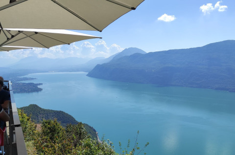Vue sur un lac paisible et des montagnes depuis une terrasse avec parasols près de Challes les Eaux, en France.