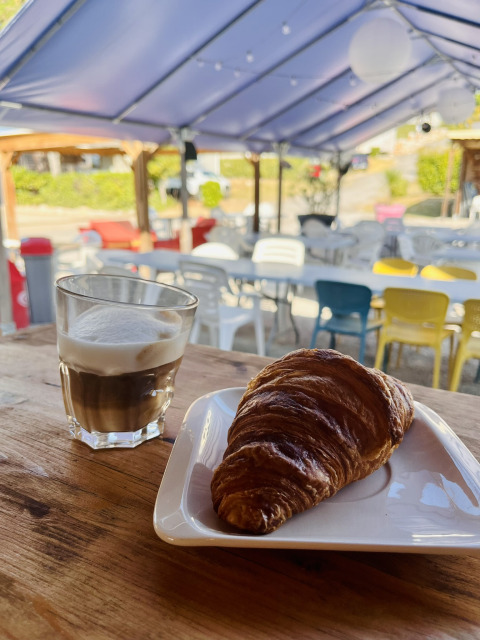 Un verre de café et un croissant sur une assiette sur une table en bois dans un café extérieur couvert.