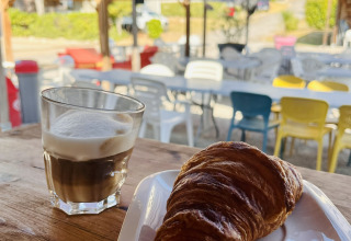 Un vaso de café y un croissant en un plato sobre una mesa de madera en una cafetería al aire libre techada.