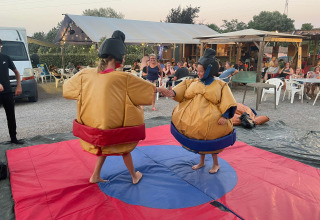 Two people in sumo suits playfully wrestle on a mat, watched by an audience at a holiday park in Provence.