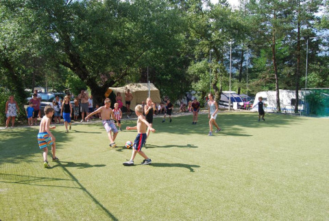 Bambini giocano a calcio nel campo di Flower Camping la Pinède, osservati da adulti e famiglie sullo sfondo.