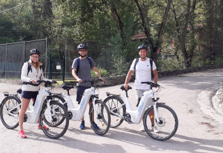 Three adults wearing bike helmets enjoy riding electric bicycles at Flower Camping la Pinède in France.