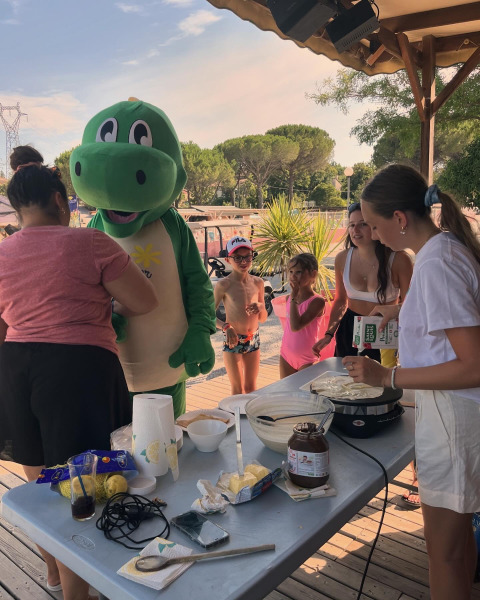Kinderen en volwassenen bakken pannenkoeken met een groene mascotte bij Flower Camping Provence Vallée, Frankrijk.