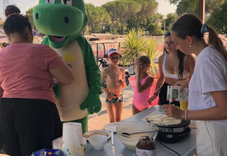 Kinder und Erwachsene genießen Pfannkuchen mit einer grünen Maskottchenfigur im Flower Camping Provence Vallée.