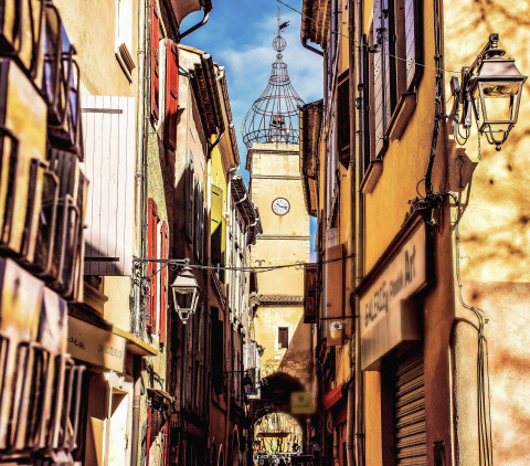 Narrow colorful street in Manosque, Provence, France, with a clock tower and bright blue sky above.