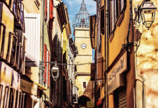 Schmale, bunte Gasse in Manosque, Provence, mit Uhrturm und typisch mediterraner Architektur.
