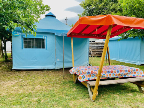 Blauwe Ecotent Lodge safaritent met picknickbank en oranje luifel op groene camping tussen bomen.