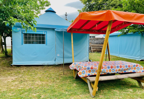 Blauwe Ecotent Lodge safaritent met picknickbank en oranje luifel op groene camping tussen bomen.