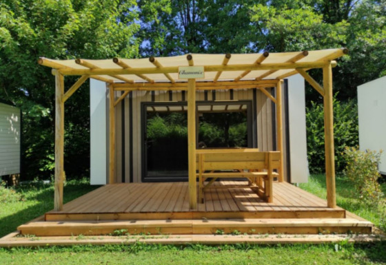 A modern glamping cabin with a wooden deck, bench seating, and pergola, surrounded by lush greenery.