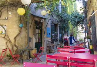 Terraza de cafetería al aire libre con mesas y sillas rosas, vegetación y ambiente de parque vacacional francés.