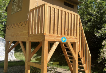 Casa sull’albero Avonturiers rialzata con terrazza in legno, mobili verdi e alberi circostanti.