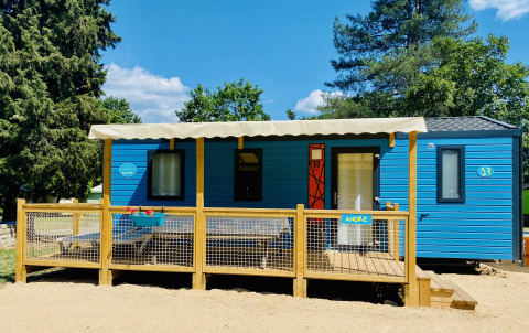Blauwe Comfort Mobile Home met drie slaapkamers, groot houten terras en luifel in groene, zonnige omgeving.