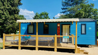 Blaues Comfort Mobile Home mit drei Schlafzimmern, Terrasse und Markise in grüner, sonniger Umgebung.
