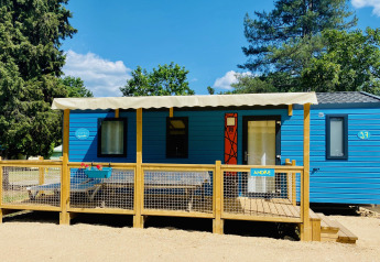 Mobil-home Comfort bleu trois chambres avec terrasse en bois et auvent, situé dans un cadre verdoyant et ensoleillé.