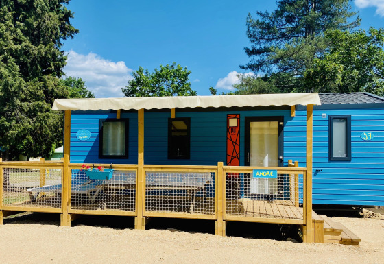 Blauwe Comfort Mobile Home met drie slaapkamers, groot houten terras en luifel in groene, zonnige omgeving.