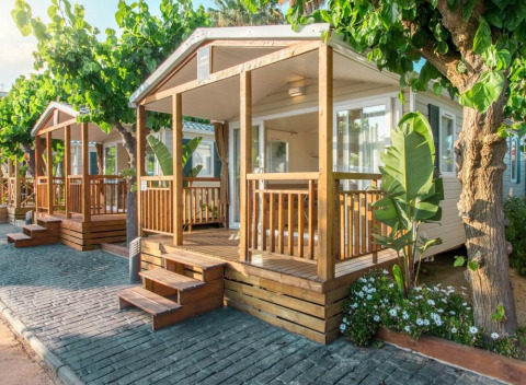 Comfort Mobile Home met twee slaapkamers, houten terras en veel groen onder schaduwrijke bomen.