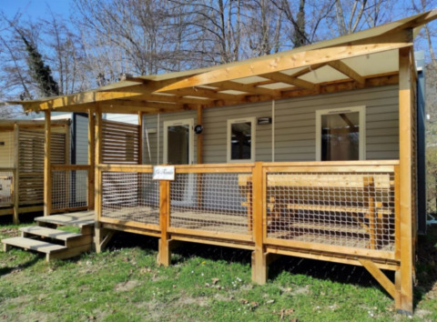 Stacaravan CONFORT 30m², moderne chalet met overdekte veranda en houten hek in een groene omgeving.