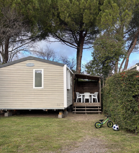Mobile Home ECO Lodgia con veranda coperta, sedie bianche, bici verde da bambino e pallone da calcio.