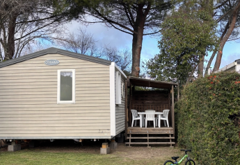 Mobilhytte ECO Lodgia med terrasse, bord og stole, grøn børnecykel og fodbold på grøn græsplæne.