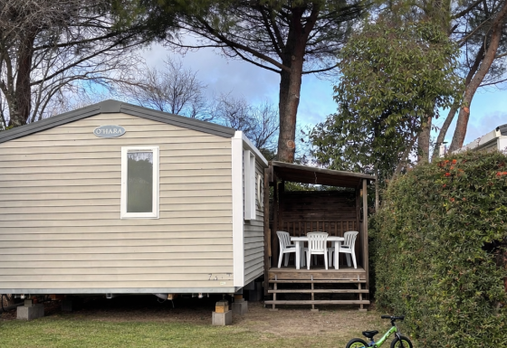 Mobile Home ECO Lodgia con terraza techada, sillas blancas, bicicleta verde infantil y balón de fútbol.