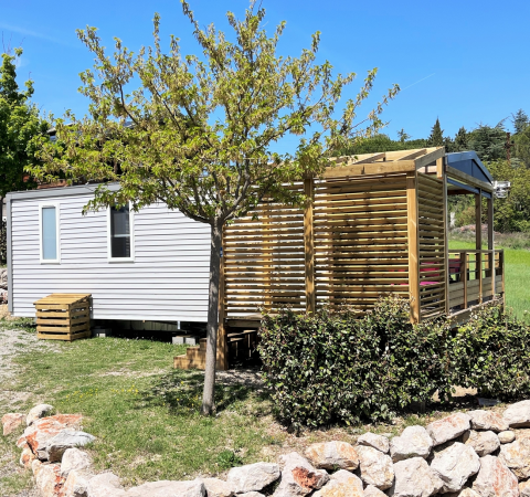Mobile Home Living Lodge avec mobil-home, terrasse boisée, verdure et ciel bleu ensoleillé à l'arrière-plan.