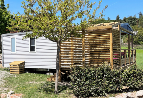 Mobile Home Living Lodge avec mobil-home, terrasse boisée, verdure et ciel bleu ensoleillé à l'arrière-plan.