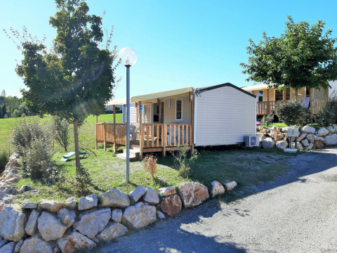 Vista esterna del Mobile Home Tribu con veranda in legno, piante verdi, rocce e cielo soleggiato.