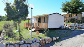 Vista esterna del Mobile Home Tribu con veranda in legno, piante verdi, rocce e cielo soleggiato.