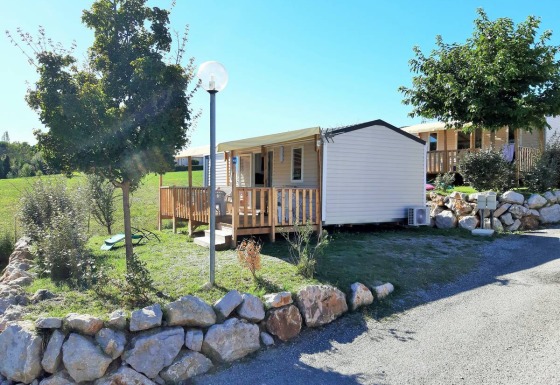 Außenansicht des Mobile Home Tribu mit Holzveranda, Steinmauern, Bäumen und sonnigem Himmel.