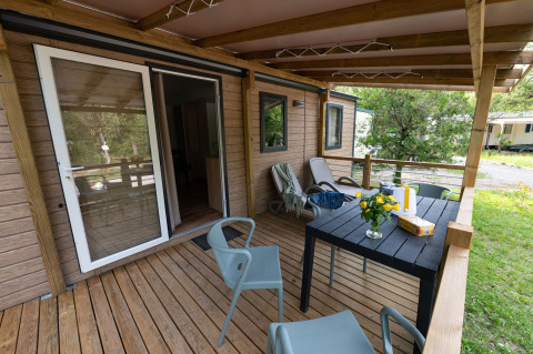 Wooden porch of a lodge with chairs, table, loungers, and flowers, surrounded by green nature in summer.