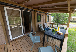 Wooden porch of a lodge with chairs, table, loungers, and flowers, surrounded by green nature in summer.