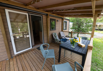 Træterrasse ved en lodge med borde, stole, liggestole og blomster, omgivet af grøn natur om sommeren.