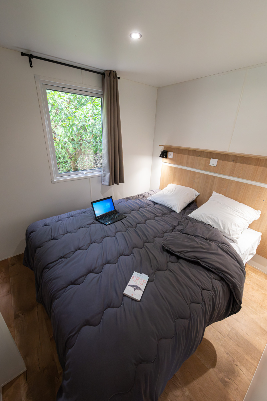 Knusse lodge slaapkamer met tweepersoonsbed, laptop, boek op het dekbed en raam met uitzicht op bomen.