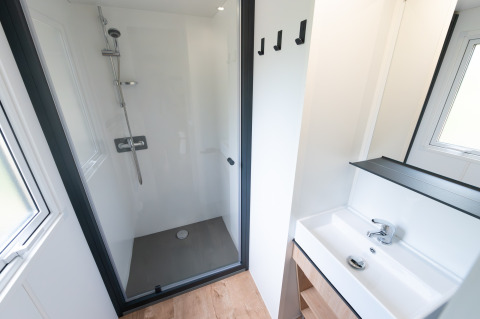 Salle de bain de lodge moderne avec douche vitrée, lavabo blanc, miroir, sol bois et fenêtre lumineuse.