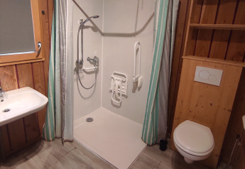 Baño accesible con ducha a ras de suelo, barras de apoyo, asiento plegable, lavabo y WC de pared.