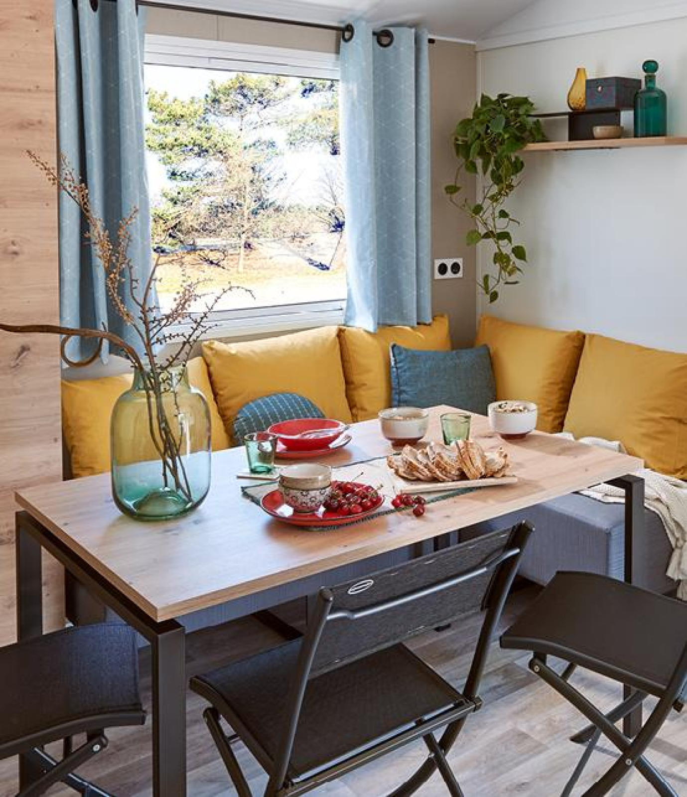 Eettafel met gele kussens, eten en raam in Comfort Mobile home bij Camping Paradis Family des Issoux.