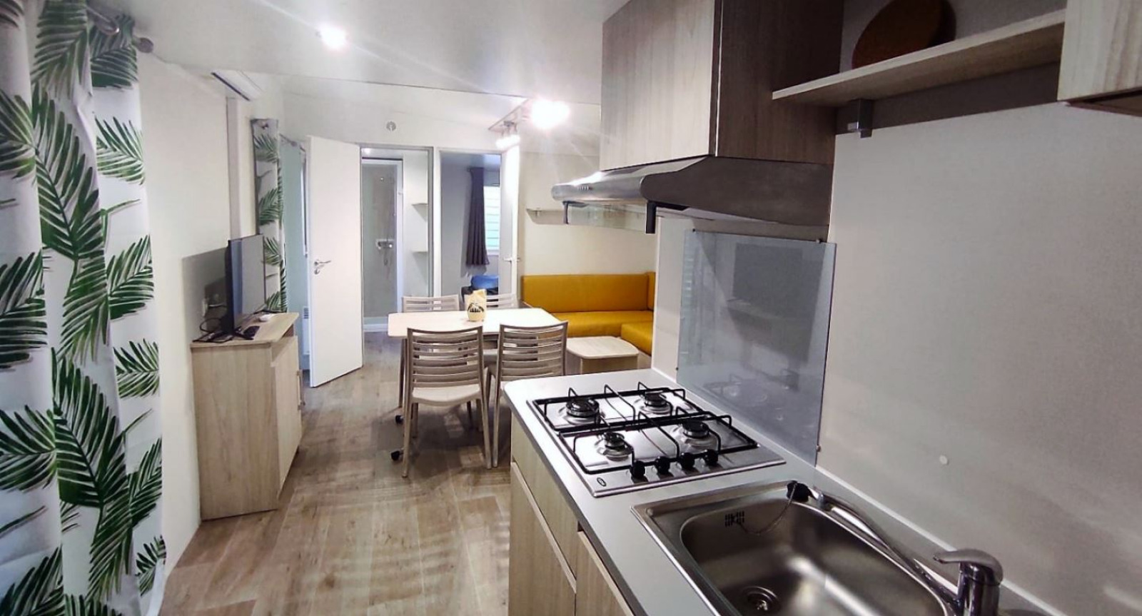 Moderne glampingaccommodatie met kitchenette, eettafel, gele bank en gordijnen met bladerpatroon.