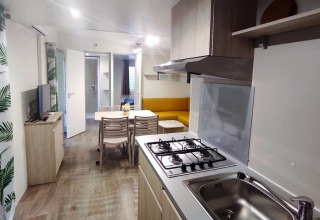 Moderne Glamping-Unterkunft mit Küchenzeile, Esstisch, Sofa, TV und Vorhängen mit Blättermuster.