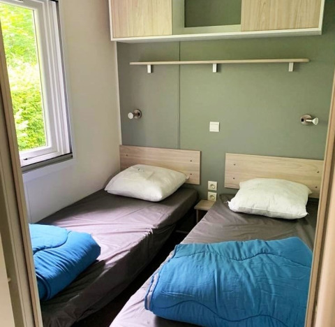 Deux lits simples avec draps gris, couettes bleues et oreillers blancs dans une chambre de glamping.