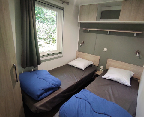 Zwei Einzelbetten mit blauer Bettdecke und großem Fenster in der Stacaravan CONFORT im Camping Le Savoy, Frankreich.