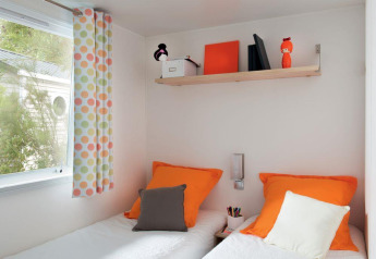 Moderne og farverig hytte med to senge, orange puder, hylde og farverige gardiner på Flower Camping Provence Vallée.