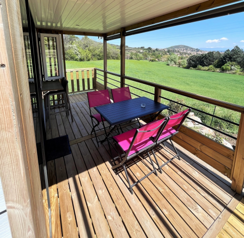 Covered wooden terrace with pink chairs and a black table at Mobile Home «Living» in Flower Camping Provence Vallée, France.