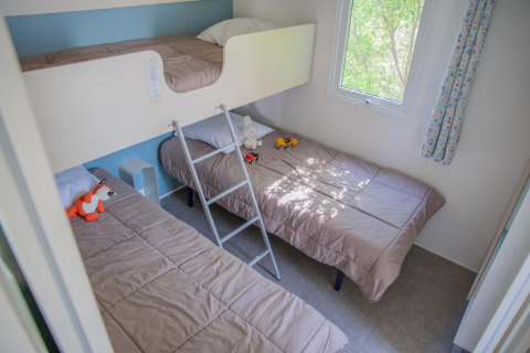 Cozy kids’ room with bunk bed, two single beds, toys on beds, and sunlit window overlooking green trees.