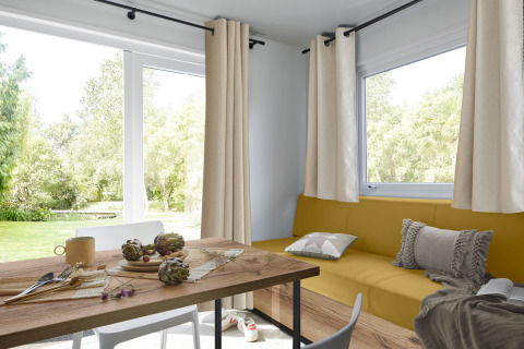 Bright living space in a Premium Mobile Home with yellow sofa, dining table, and garden view in France.