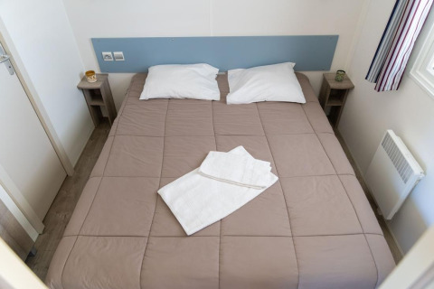 Chambre de Premium Mobile Home avec lit double, deux oreillers, serviettes et tables de nuit au Flower Camping Provence Vallée.