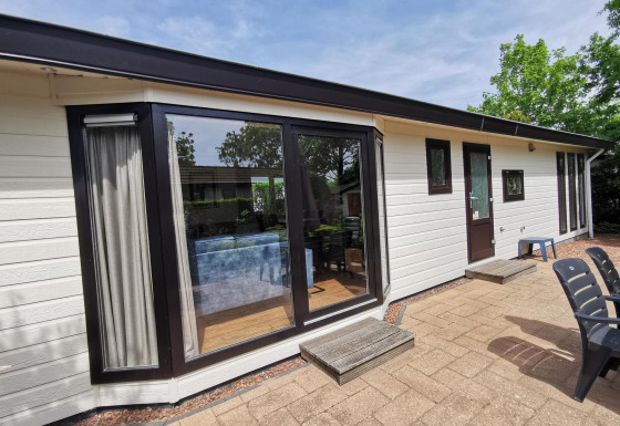Outdoor photo of a modern cabin named Comfort featuring large glass windows, patio, and outdoor chairs in sun.