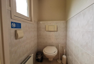 Bathroom with wall-mounted toilet, toilet brush, trash bin, and window at the 'Comfort' cabin in De Woudhoeve.
