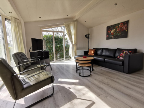 Bright living room in Comfort cabin at De Woudhoeve, Netherlands, with black sofa, chairs, TV and large windows.