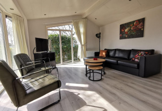 Bright living room in Comfort cabin at De Woudhoeve, Netherlands, with black sofa, chairs, TV and large windows.
