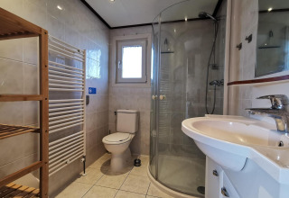Modern bathroom with glass shower, toilet, sink, wooden shelf and window in Comfort cabin at De Woudhoeve.